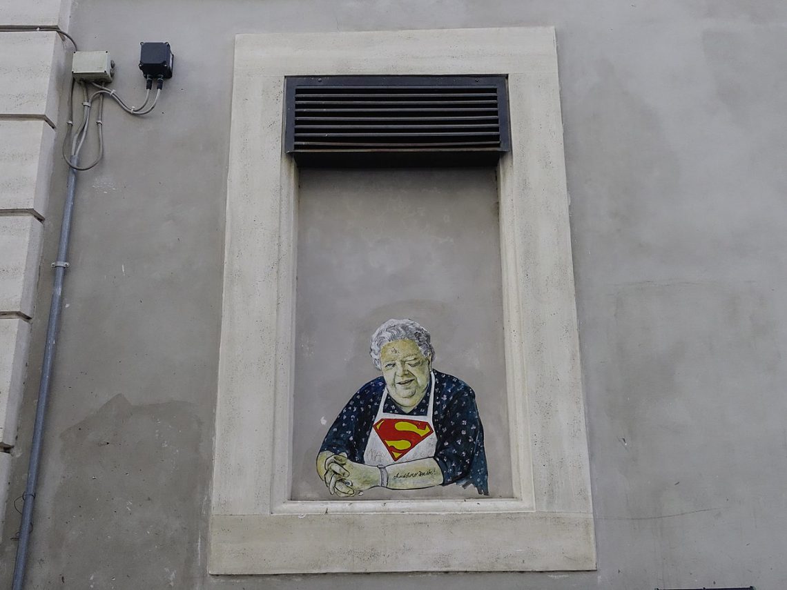 Su un'asse che chiude una finestra incastonata in un muro di cemento grigio e contornata da una specie di cornice di cemento più chiaro c'è un murale che ritrae la sora Lella (l'attrice Elena Fabrizi) come fosse seduta alla finestra, con le braccia appoggiate sul davanzale, mentre sorridendo fa l'occhiolino con l'occhio sinistro. Hai i capelli bianchi, indossa una veste blu a motivi bianchi e, sopra quella, un grembiule bianco con il simbolo di Superman sul petto.