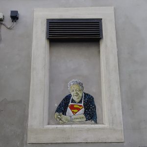 Su un'asse che chiude una finestra incastonata in un muro di cemento grigio e contornata da una specie di cornice di cemento più chiaro c'è un murale che ritrae la sora Lella (l'attrice Elena Fabrizi) come fosse seduta alla finestra, con le braccia appoggiate sul davanzale, mentre sorridendo fa l'occhiolino con l'occhio sinistro. Hai i capelli bianchi, indossa una veste blu a motivi bianchi e, sopra quella, un grembiule bianco con il simbolo di Superman sul petto.