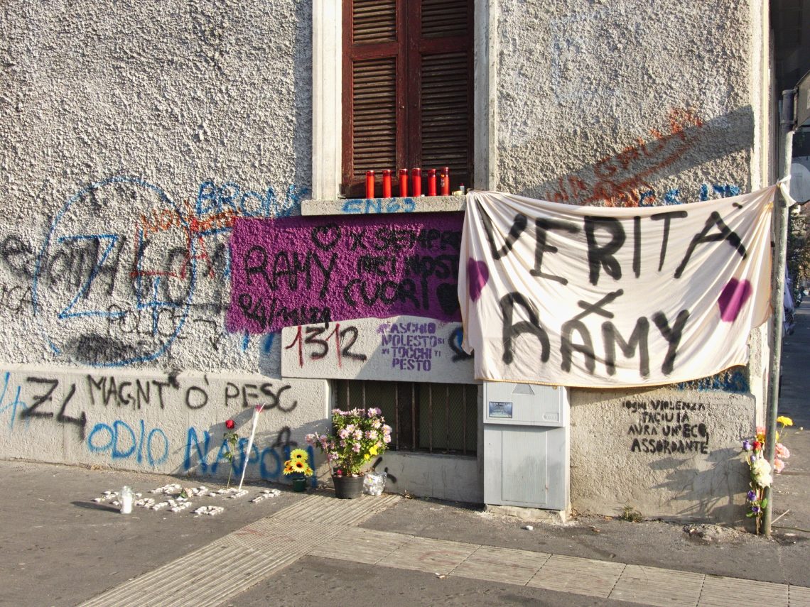 Una foto a colori, in formato orizzontale (4:3), che ritrae un muro d'angolo della parte inferiore - dal suolo fino al primo piano - di una casa. Sul muro c'è una scritta, "Ramy per sempre nei nostri cuori"; ce n'è un'altra, "Verità per Ramy", su uno striscione bianco appeso a sinistra a un fermaglio all'angolo inferiore destro di una finestra chiusa, e a destra a un palo segnaletico. Sul davanzale della finestra ci sono inoltre sei ceri rossi. In basso, sul marciapiedi, ci sono fiori singoli (due rose rosse), due vasetti e un mazzo di fiori vari. Davanti alle rose, sull'asfalto, ci sono tante candeline bianche, spente.