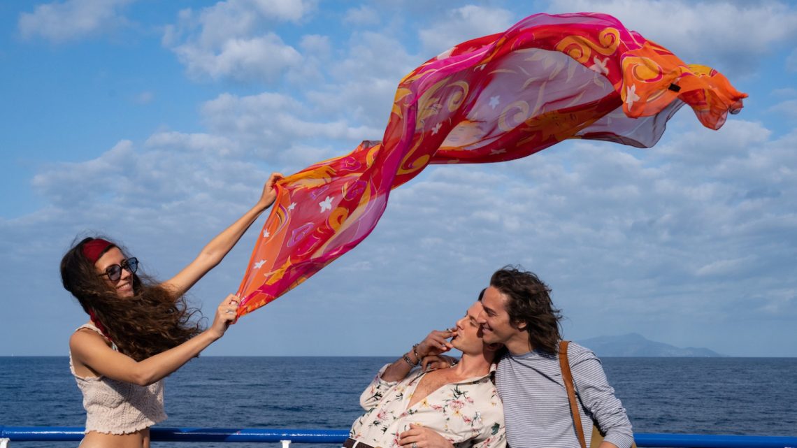 Un fotogramma dal film "Parthenope" di Paolo Sorrentino. Sullo sfondo di un mare blu e un cielo azzurro con nuvolette bianche, una ragazza, in piedi a sinistra nell'inquadratura, tiene con le braccia alzate in obliquo verso destra un ampio velo rosso e arancione, decorato con motivi astratti, che garrisce nel vento sopra le teste di due ragazzi sorridenti, i quali, di profilo, sulla destra dell'inquadratura, guardano la ragazza, il primo fumando una sigaretta, col busto piegato all'indietro e la nuca poggiata sulla spalla del secondo.