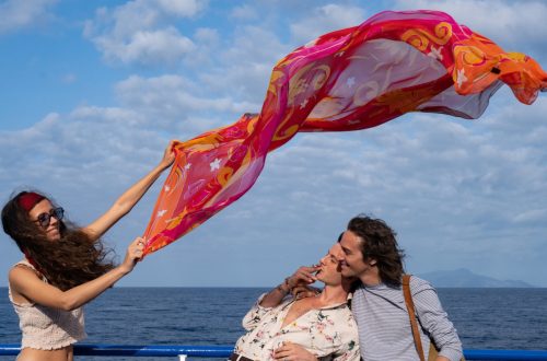 Un fotogramma dal film "Parthenope" di Paolo Sorrentino. Sullo sfondo di un mare blu e un cielo azzurro con nuvolette bianche, una ragazza, in piedi a sinistra nell'inquadratura, tiene con le braccia alzate in obliquo verso destra un ampio velo rosso e arancione, decorato con motivi astratti, che garrisce nel vento sopra le teste di due ragazzi sorridenti, i quali, di profilo, sulla destra dell'inquadratura, guardano la ragazza, il primo fumando una sigaretta, col busto piegato all'indietro e la nuca poggiata sulla spalla del secondo.