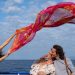 Un fotogramma dal film "Parthenope" di Paolo Sorrentino. Sullo sfondo di un mare blu e un cielo azzurro con nuvolette bianche, una ragazza, in piedi a sinistra nell'inquadratura, tiene con le braccia alzate in obliquo verso destra un ampio velo rosso e arancione, decorato con motivi astratti, che garrisce nel vento sopra le teste di due ragazzi sorridenti, i quali, di profilo, sulla destra dell'inquadratura, guardano la ragazza, il primo fumando una sigaretta, col busto piegato all'indietro e la nuca poggiata sulla spalla del secondo.