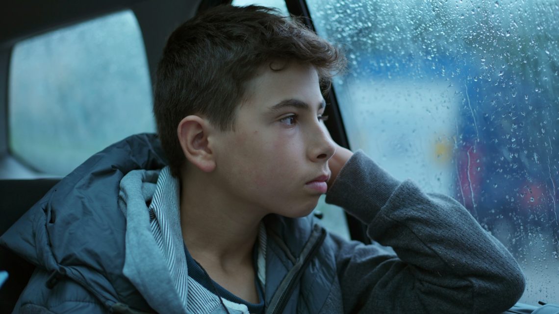 Una foto che ritrae il mezzobusto di un ragazzo seduto sul sedile posteriore di un'auto mentre guarda attraverso il finestrino alla sua sinistra, ricoperto all'esterno di gocce e rivoletti di pioggia, un paesaggio piovoso fuori fuoco. Il ragazzo è di profilo, con il volto girato verso la sua sinistra e il gomito del braccio sinistro appoggiato alla portiera. Ha un'espressione pensierosa, molto seria, un po' scontenta.