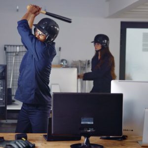 Una foto a colori, in formato orizzontale (16:9), che ritrae un uomo e una donna in una “stanza della rabbia”. Entrambi indossano camicia e pantaloni blu, e un casco da baseball sulla testa. L’uomo brandisce una mazza da baseball con cui è in procinto di sferrare un colpo a un monitor da computer.