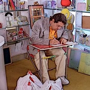 Un fotogramma dal film “Da grande” (1987), di Franco Amurri, con Renato Pozzetto. Il personaggio interpretato da Pozzetto, un bambino che all’improvviso si ritrova nel suo corpo da adulto, è seduto a un piccolo banco rosso per bambini, su una piccola sedia rossa, impegnato a scrivere con una penna su un foglio, all’interno di una cartoleria.