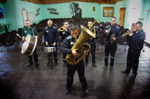Una foto a colori, in formato orizzontale, che ritrae una banda musicale formata da sette carcerati, in piedi con vari strumenti davanti a una platea di sedie vuote. In primo piano c'è un uomo con un trombone, gli altri sei sono disposti in fila alle sue spalle.