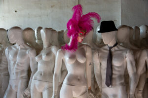 Una foto a colori, in formato orizzontale, che ritrae tanti manichini senza volto, verniciati di bianco, con la vernice un po' scrostata, graffiata o sporca in alcune parti, all'interno di una stanza dalle pareti intonacate di bianco sporco. Al centro ce n'è uno dalle fattezze femminili che indossa una maschera veneziana e un copricapo di lunghe piume fuxia; al suo fianco, sulla destra dell'inquadratura, ce n'è uno dalle fattezze maschili con un cappello nero in testa e una cravatta nera al collo.