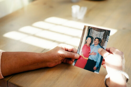 Una foto a colori, in formato orizzontale, che ritrae due mani femminili poggiate su un tavolo che reggono una foto che a sua volta ritrae una donna e un uomo che si abbracciano sorridendo e guardando verso l'obiettivo.