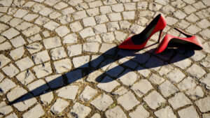 Una foto a colori, in formato orizzontale, che ritrae in alto a destra un paio di scarpe rosse con tacco da donna appoggiate su un selciato di sanpietrini che occupa tutto il resto dell'inquadratura. La scarpa destra è in piedi, la sinistra è appoggiata sul fianco destro. Entrambe proiettano sul selciato, da destra a sinistra, una lunga ombra scura.