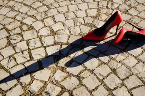 Una foto a colori, in formato orizzontale, che ritrae in alto a destra un paio di scarpe rosse con tacco da donna appoggiate su un selciato di sanpietrini che occupa tutto il resto dell'inquadratura. La scarpa destra è in piedi, la sinistra è appoggiata sul fianco destro. Entrambe proiettano sul selciato, da destra a sinistra, una lunga ombra scura.