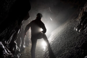Una foto a colori, in formato orizzontale, che ritrae al centro di un cunicolo sotterraneo uno speleologo di spalle che illumina la strada davanti a sé con una torcia.