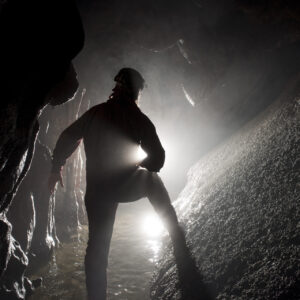 Una foto a colori, in formato orizzontale, che ritrae al centro di un cunicolo sotterraneo uno speleologo di spalle che illumina la strada davanti a sé con una torcia.