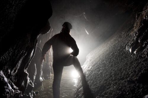 Una foto a colori, in formato orizzontale, che ritrae al centro di un cunicolo sotterraneo uno speleologo di spalle che illumina la strada davanti a sé con una torcia.