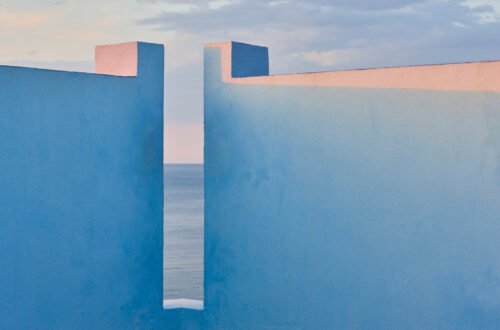Un dipinto che raffigura un angolo formato da due muri bianchi illuminati da una luce d'alba o tramonto. In corrispondenza dell'angolo le mura si aprono sulla vista di un mare lontano, azzurro e placido fino all'orizzonte, sopra il quale sta un cielo di colore azzurro più chiaro, con qualche nuvola grigia che si allunga in orizzontale a destra.