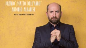 Locandina della settima edizione del premio "Matto dell'anno". C'è una foto a colori, in formato verticale, che ritrae Antonio Albanese in piedi davanti a un muro intonacato di color giallo ocra. Albanese è ritratto dalle anche in su, indossa un completo nero, ha le mani unite a pugno tenute davanti al petto, un'espressione seria di gratitudine e combattività sul volto. In alto a sinistra c'è una scritta bianca: «7^ edizione - premio "Matto dell'anno" - Antonio Albanese»; sotto, in piccolo e in nero: «Musica di Marina Notaro e Alessandro Acri». Nell'angolo in basso a sinistra dell'inquadratura ci sono due loghi affiancati: "MAO" e "Fondazione Lighea onlus", quest'ultimo con la grafica in azzurro che raffigura la sirena Lighea di profilo con le mani alzate davanti al viso, come se cantasse a gran voce, sopra la scritta già citata, in nero.