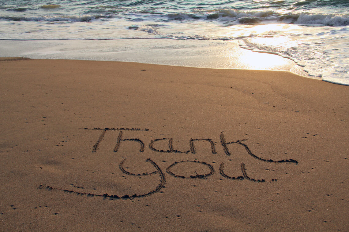 Una foto a colori, in formato orizzontale, che ritrae una scritta "Thank you" tracciata con un dito nella sabbia della battigia su una spiaggia davanti al mare.