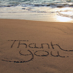 Una foto a colori, in formato orizzontale, che ritrae una scritta "Thank you" tracciata con un dito nella sabbia della battigia su una spiaggia davanti al mare.