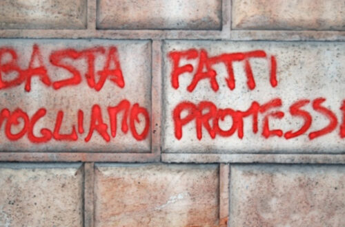 Una foto a colori, orizzontale, che ritrae un muro con una scritta a spray rosso: "Basta fatti vogliamo promesse".