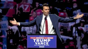 Una foto che ritrae Charlie Kirk su un podio con le scritte "Text Arizona to 88022 - Trump - Vance - Make America great again! - 2024", davanti a una platea con tanta gente, a braccia allargate, durante un discorso di sostegno a Trump per le elezioni 2024.