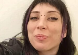 Un selfie in formato orizzontale di Cinzia Pinna, sorridente, con due ciocche di capelli viola a incorniciare il bel viso, un piercing sul labbro inferiore e uno all'attaccatura del naso.