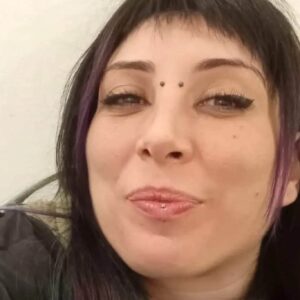 Un selfie in formato orizzontale di Cinzia Pinna, sorridente, con due ciocche di capelli viola a incorniciare il bel viso, un piercing sul labbro inferiore e uno all'attaccatura del naso.