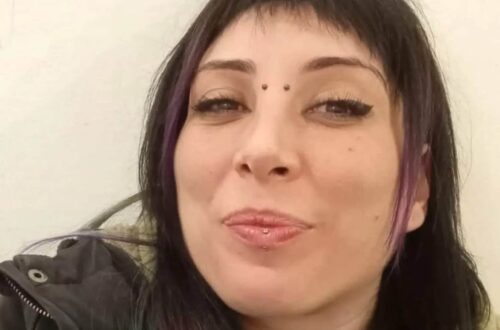 Un selfie in formato orizzontale di Cinzia Pinna, sorridente, con due ciocche di capelli viola a incorniciare il bel viso, un piercing sul labbro inferiore e uno all'attaccatura del naso.