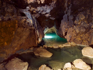 Una foto a colori, in formato orizzontale, che ritrae alcuni piccoli laghetti d'acqua trasparente, di colore verde-azzurro, in una caverna parizlalmente illuminata dall'alto dalla luce del sole che entra attraverso dei buchi sul suo soffitto.