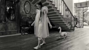 Un fotogramma dal film "Povere creature", di Yorgos Lathimos. La ragazza protagonista, in vestaglia, cammina a piedi scalzi su un parquet all'interno di una grande casa, seguita a pochi metri da uno strano essere con quattro zampe che sbucano da un tronco orizzontale (potrebbe essere il corpo di un cane glabro), e un lungo, flessuoso collo bianco con testa e becco giallo da papera.