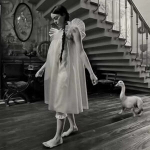 Un fotogramma dal film "Povere creature", di Yorgos Lathimos. La ragazza protagonista, in vestaglia, cammina a piedi scalzi su un parquet all'interno di una grande casa, seguita a pochi metri da uno strano essere con quattro zampe che sbucano da un tronco orizzontale (potrebbe essere il corpo di un cane glabro), e un lungo, flessuoso collo bianco con testa e becco giallo da papera.