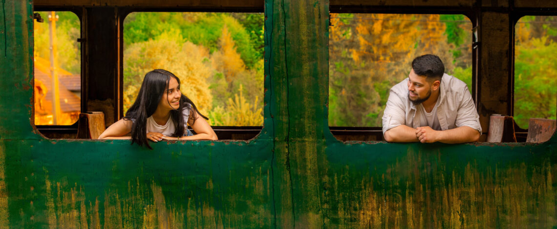 Una foto a colori, in formato orizzontale, che ritrae una giovane donna e un giovane uomo, forse una coppia, affacciati ciascuno a una finestra diversa, senza più vetri, di un vagone ferroviario dismesso, fermo davanti a una boscaglia dai colori autunnali. I due si guardano sorridendo.