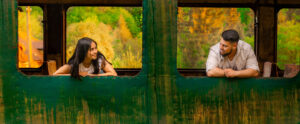 Una foto a colori, in formato orizzontale, che ritrae una giovane donna e un giovane uomo, forse una coppia, affacciati ciascuno a una finestra diversa, senza più vetri, di un vagone ferroviario dismesso, fermo davanti a una boscaglia dai colori autunnali. I due si guardano sorridendo.