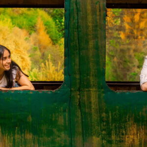 Una foto a colori, in formato orizzontale, che ritrae una giovane donna e un giovane uomo, forse una coppia, affacciati ciascuno a una finestra diversa, senza più vetri, di un vagone ferroviario dismesso, fermo davanti a una boscaglia dai colori autunnali. I due si guardano sorridendo.