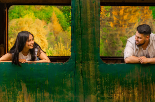Una foto a colori, in formato orizzontale, che ritrae una giovane donna e un giovane uomo, forse una coppia, affacciati ciascuno a una finestra diversa, senza più vetri, di un vagone ferroviario dismesso, fermo davanti a una boscaglia dai colori autunnali. I due si guardano sorridendo.