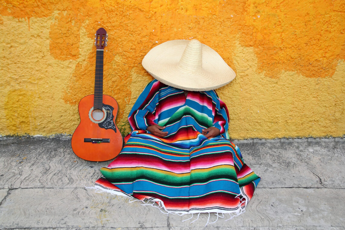 Una foto a colori, in formato orizzontale, che ritrae al centro un uomo che fa la siesta seduto per terra, su un marciapiedi grigio, con la schiena appoggiata a un muro giallo. L'uomo è completamente avvolto in un poncho colorato a strisce orizzontali da cui fuoriescono solo le mani, appoggiate in grembo, e sulla testa indossa un sombrero inclinato in avanti a coprire completamente anche il viso. Al suo fianco, a sinistra, appoggiata in verticale sul marciapiedi e contro il muro giallo, c'è una chitarra classica con la cassa arancione.