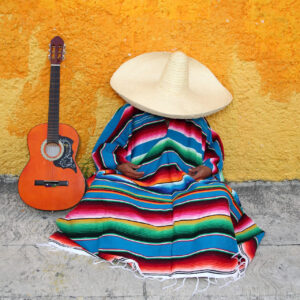 Una foto a colori, in formato orizzontale, che ritrae al centro un uomo che fa la siesta seduto per terra, su un marciapiedi grigio, con la schiena appoggiata a un muro giallo. L'uomo è completamente avvolto in un poncho colorato a strisce orizzontali da cui fuoriescono solo le mani, appoggiate in grembo, e sulla testa indossa un sombrero inclinato in avanti a coprire completamente anche il viso. Al suo fianco, a sinistra, appoggiata in verticale sul marciapiedi e contro il muro giallo, c'è una chitarra classica con la cassa arancione.