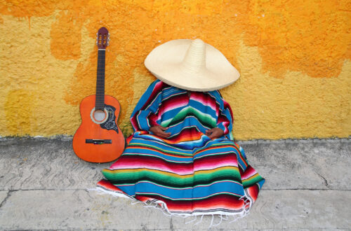 Una foto a colori, in formato orizzontale, che ritrae al centro un uomo che fa la siesta seduto per terra, su un marciapiedi grigio, con la schiena appoggiata a un muro giallo. L'uomo è completamente avvolto in un poncho colorato a strisce orizzontali da cui fuoriescono solo le mani, appoggiate in grembo, e sulla testa indossa un sombrero inclinato in avanti a coprire completamente anche il viso. Al suo fianco, a sinistra, appoggiata in verticale sul marciapiedi e contro il muro giallo, c'è una chitarra classica con la cassa arancione.