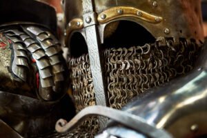 Una foto a colori, in formato orizzontale, che ritrae alcuni particolari di un'armatura medievale metallica: un elmo e il guanto della mano destra appoggiato allo stesso, in una posizione che, se nell'armatura ci fosse qualcuno, potrebbe apparire pensosa.