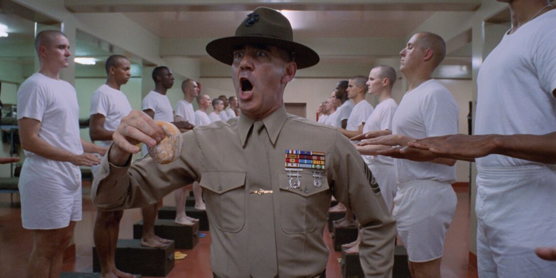 Un fotogramma dal film "Full Metal Jacket", di Stanley Kubrick. Al centro dell'inquadratura, in piedi, è ritratto dalla cintola in su un istruttore militare urlante, con espressione da invasato e un krpfen nella mano destra, mentre incede attraverso due file, una a sinistra e una a destra, di soldati in piedi di profilo, in maglietta e boxer bianchi, ciascuno con le mani tese davanti a sé, pronte a ricevere la punizione dell'istruttore.