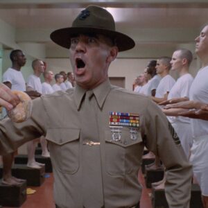 Un fotogramma dal film "Full Metal Jacket", di Stanley Kubrick. Al centro dell'inquadratura, in piedi, è ritratto dalla cintola in su un istruttore militare urlante, con espressione da invasato e un krpfen nella mano destra, mentre incede attraverso due file, una a sinistra e una a destra, di soldati in piedi di profilo, in maglietta e boxer bianchi, ciascuno con le mani tese davanti a sé, pronte a ricevere la punizione dell'istruttore.