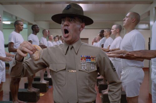 Un fotogramma dal film "Full Metal Jacket", di Stanley Kubrick. Al centro dell'inquadratura, in piedi, è ritratto dalla cintola in su un istruttore militare urlante, con espressione da invasato e un krpfen nella mano destra, mentre incede attraverso due file, una a sinistra e una a destra, di soldati in piedi di profilo, in maglietta e boxer bianchi, ciascuno con le mani tese davanti a sé, pronte a ricevere la punizione dell'istruttore.
