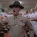Un fotogramma dal film "Full Metal Jacket", di Stanley Kubrick. Al centro dell'inquadratura, in piedi, è ritratto dalla cintola in su un istruttore militare urlante, con espressione da invasato e un krpfen nella mano destra, mentre incede attraverso due file, una a sinistra e una a destra, di soldati in piedi di profilo, in maglietta e boxer bianchi, ciascuno con le mani tese davanti a sé, pronte a ricevere la punizione dell'istruttore.