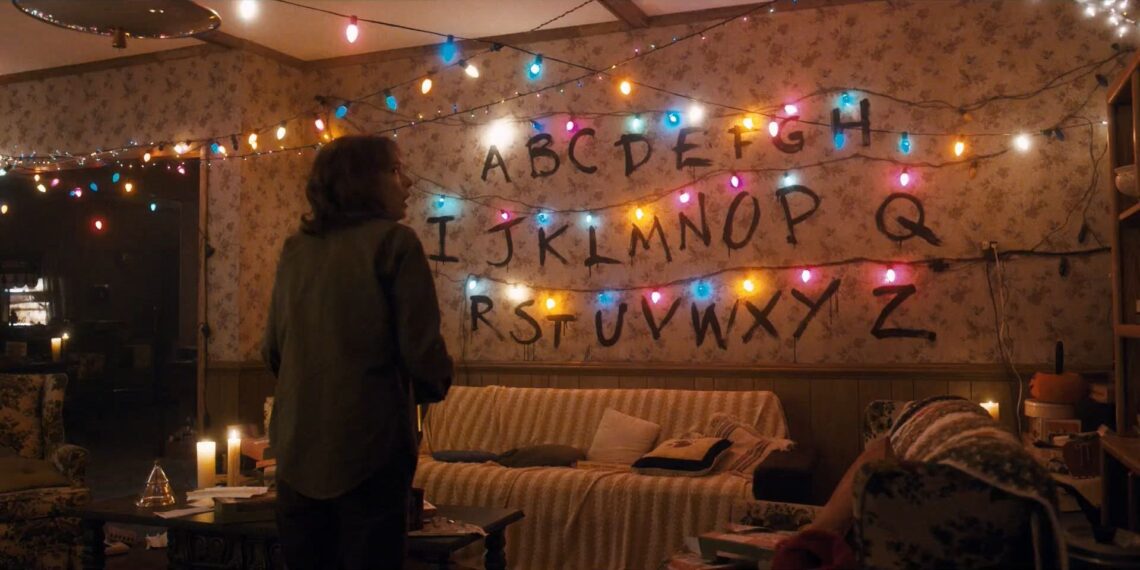 Un fotogramma dalla serie "Stranger Things". Sulla parete di fondo di un salotto illuminato da luci natalizie sono tracciate in serie tutte le lettere dell'alfabeto, sormontate ciascuna da una lucina natalizia. Al centro della stanza c'è una donna in piedi, di schiena, di profilo, con la testa girata verso destra e un' espressione spaventata sul volto.