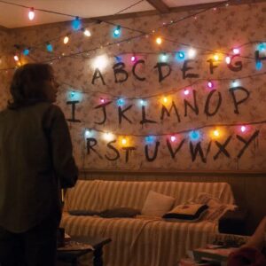 Un fotogramma dalla serie "Stranger Things". Sulla parete di fondo di un salotto illuminato da luci natalizie sono tracciate in serie tutte le lettere dell'alfabeto, sormontate ciascuna da una lucina natalizia. Al centro della stanza c'è una donna in piedi, di schiena, di profilo, con la testa girata verso destra e un' espressione spaventata sul volto.