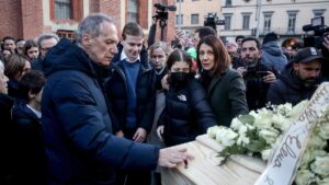 Una foto a colori, in formato orizzontale, che ritrae i famigliari di una delle ragazze morte nella tragedia di Crans Montana mentre osservano con espressioni addoloratissime la bara in cui è riposta, esposta tra fotografi e altre persone nella via di una città.