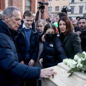 Una foto a colori, in formato orizzontale, che ritrae i famigliari di una delle ragazze morte nella tragedia di Crans Montana mentre osservano con espressioni addoloratissime la bara in cui è riposta, esposta tra fotografi e altre persone nella via di una città.