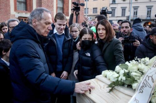 Una foto a colori, in formato orizzontale, che ritrae i famigliari di una delle ragazze morte nella tragedia di Crans Montana mentre osservano con espressioni addoloratissime la bara in cui è riposta, esposta tra fotografi e altre persone nella via di una città.