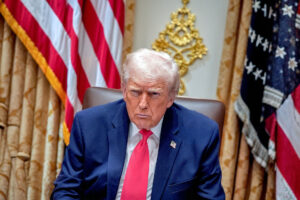 Una foto che ritrae l'attuale presidente degli Stati Uniti d'America, Donald Trump, seduto davanti a due bandiere americane, con un'espressione torva sul volto.
