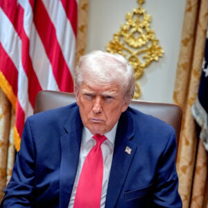 Una foto che ritrae l'attuale presidente degli Stati Uniti d'America, Donald Trump, seduto davanti a due bandiere americane, con un'espressione torva sul volto.
