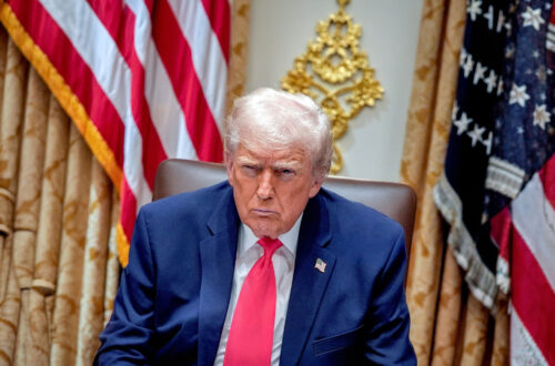 Una foto che ritrae l'attuale presidente degli Stati Uniti d'America, Donald Trump, seduto davanti a due bandiere americane, con un'espressione torva sul volto.