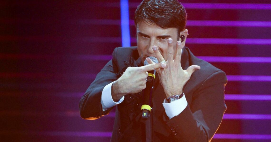 Una foto di scena dal palco del teatro Ariston durante Sanremo 2026: il vincitore, Sal Da Vinci, indica con lindice della mano destra l'anello nuziale che indossa sull'anulare della mano sinistra aperta davanti alla faccia, con un'espressione aggressiva sul volto.